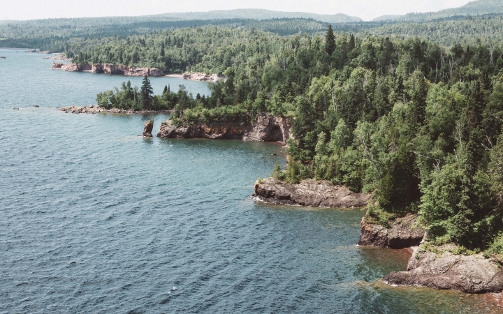 The rocky and tree-lined shore of Lake Superior frames the blue water of the lake. 
